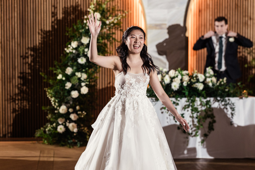Bride in a white gown waves, smiling, with floral decorations and groom in the background during Chicago Winery wedding reception