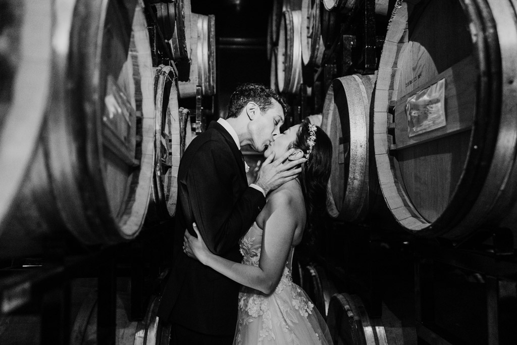 Black and white photo of newlyweds kissing between large wine barrels in the barrel room of Chicago Winery