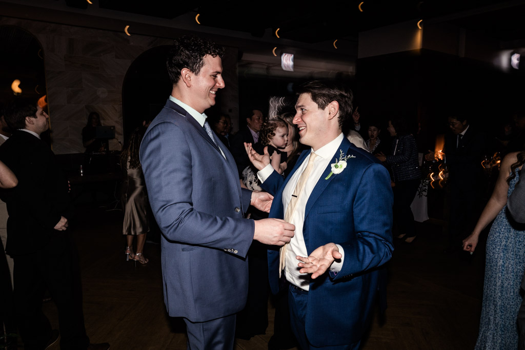 Two guests in blue suits smile and talk during wedding reception at Chicago Winery, with other people mingling in the background