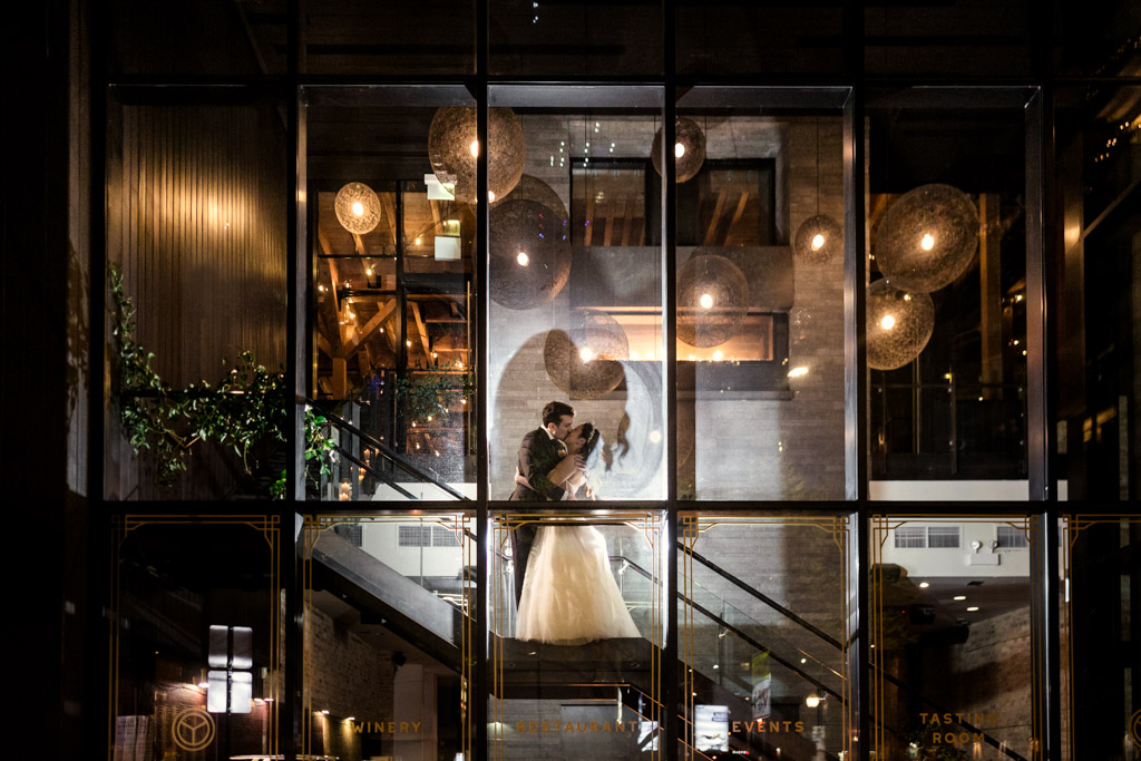 Bride and groom kiss on a staircase at Chicago Winery, seen through large glass windows at night with warm lights inside
