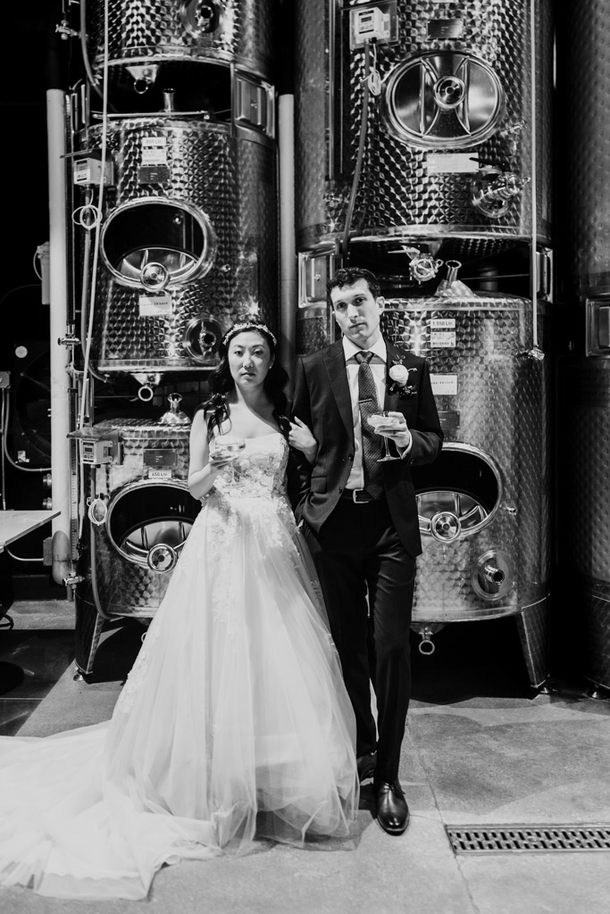 Black and white photo of bride and groom pose together in front of large metal tanks at Chicago Winery