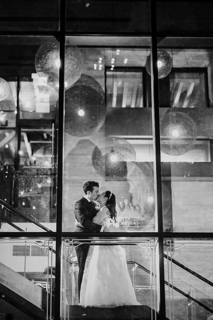 Black and white photo of bride and groom kissing on a staircase behind large glass windows at night during their Chicago Winery wedding reception