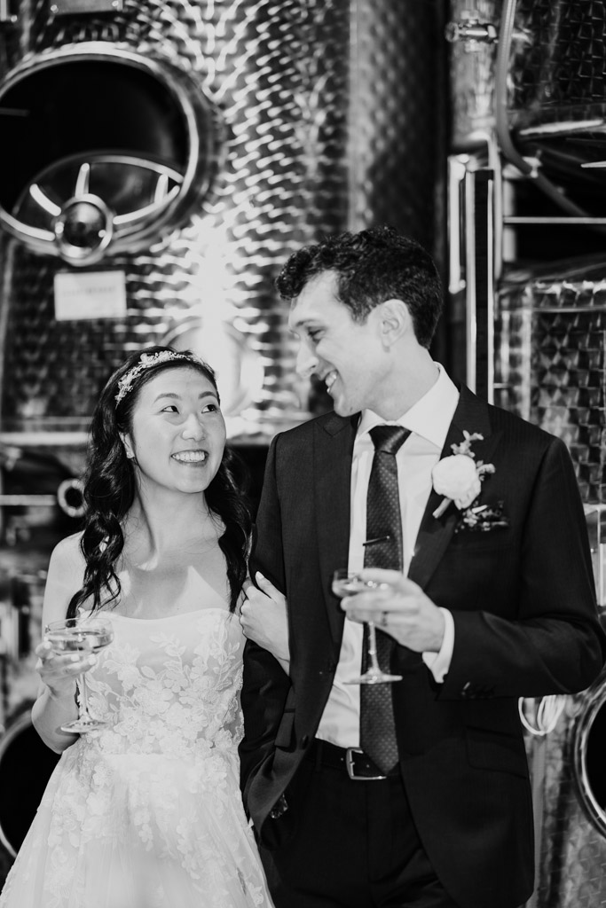 Black and white photo bride and groom smiling at each other, holding drinks, standing in front of shiny metal tanks at Chicago Winery