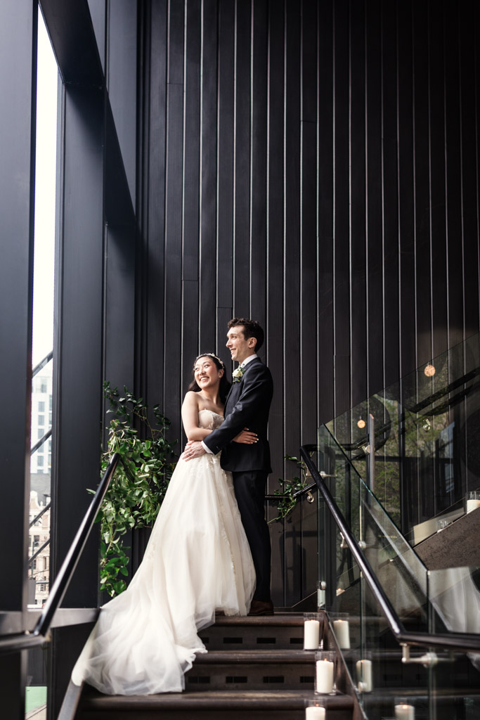 Bride and groom stand smiling together on a modern staircase at Chicago Winery, holding each other and looking out a window