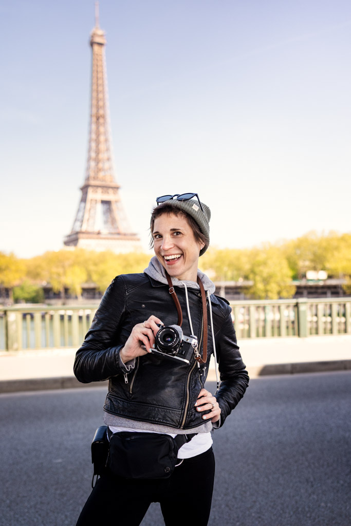 Smiling person with a camera poses on a street with the Eiffel Tower in the background, capturing memories to remember their travel dates.