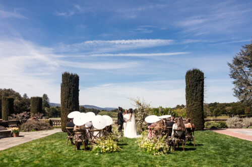 Bride and groom stand at an outdoor wedding ceremony with guests seated under white parasols.
