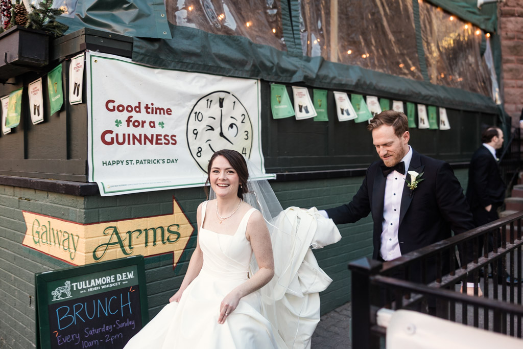 Bride and groom in formal attire smile outside Galway Arms Pub before their Cafe Brauer wedding reception