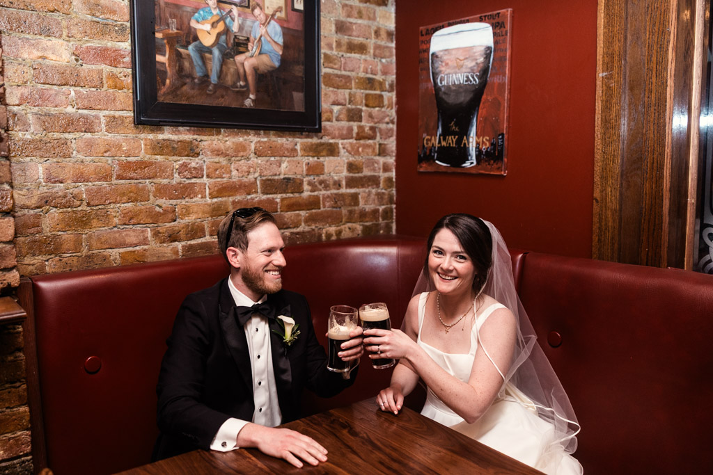 Bride and groom in wedding attire clink glasses of beer at Galway Arms after their wedding ceremony