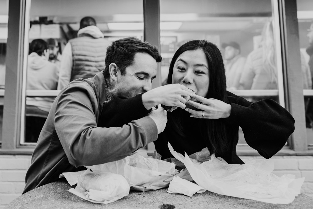 Fun black and white photo of man and woman sharing food at a table outside Johnnie's Beef holding sandwiches with both hands during their Oak Park engagement session