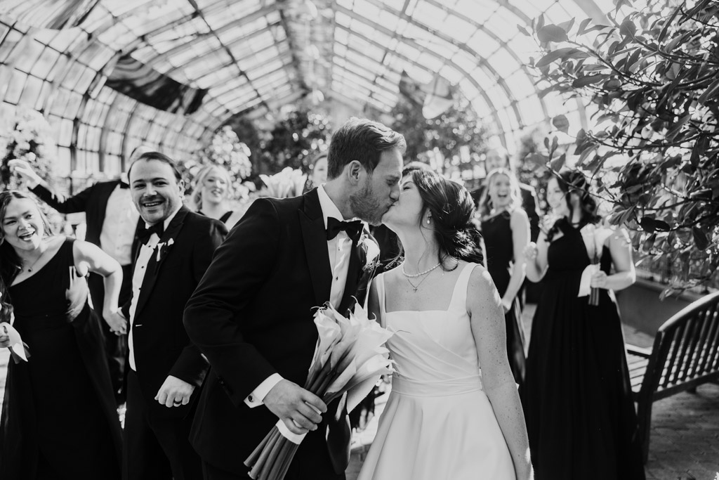 Black and white photo of bride and groom kissing while their wedding party cheers behind them inside Lincoln Park Conservatory in Chicago