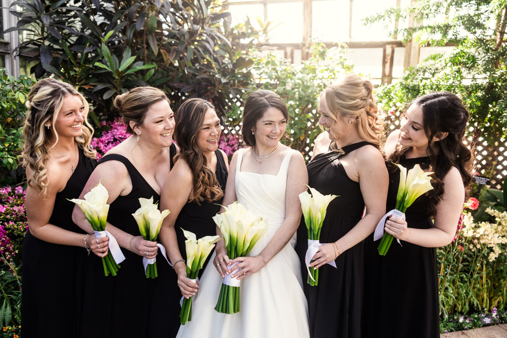 Portrait of bride in a white dress with five bridesmaids in black dresses, all holding white calla lilies and smiling together inside Lincoln Park Conservatory in Chicago