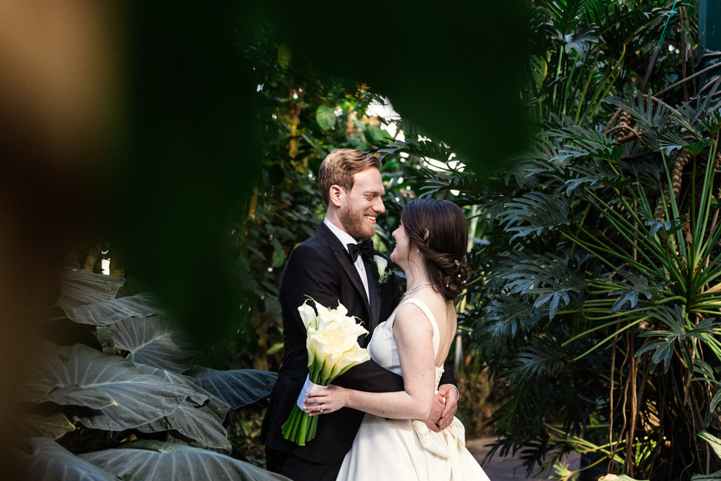 Candid photo of bride and groom smiling at each other amid lush green foliage in side Lincoln Park Conservatory in Chicago