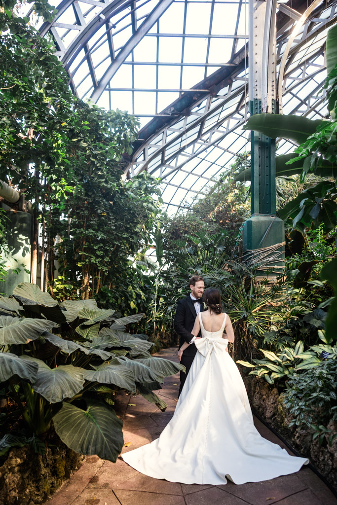 Bride and groom walk along a path through lush green plants inside a greenhouse at Lincoln Park Conservatory in Chicago