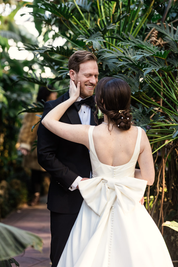 Bride touches groom's face lovinglyas they stand surround by lush green foliage inside Lincoln Park Conservatory in Chicago