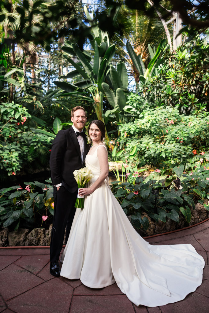 Portrait of happy bride and groom in front of lush plants and flowers inside Lincoln Park Conservatory in Chicago