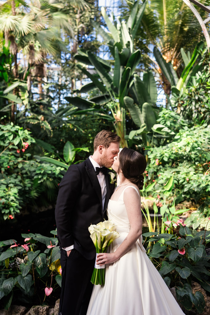 A bride and groom kiss inside Lincoln Park Conservatory in Chicago, the bride holding a bouquet of white flowers