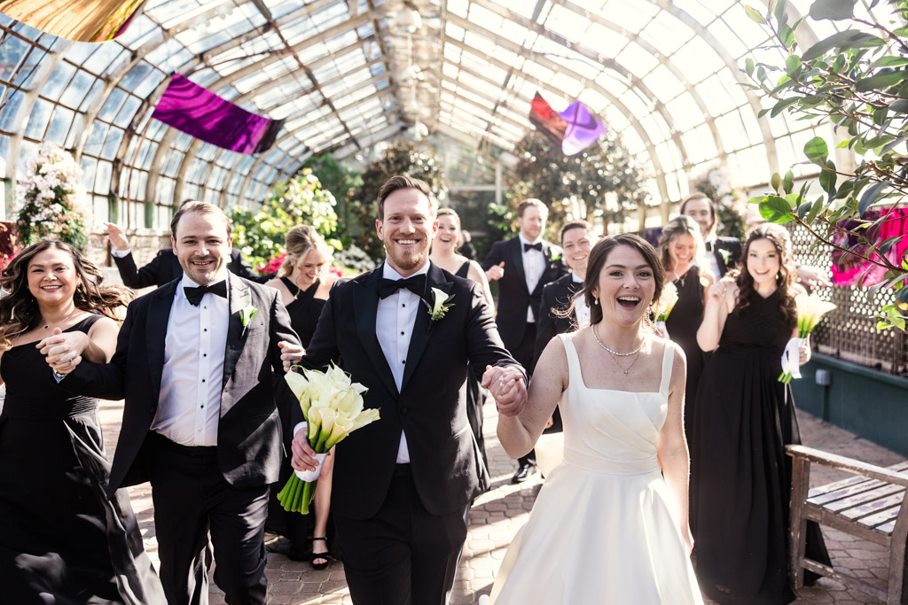 Joyful bride and groom walk hand in hand with their wedding party through a greenhouse at Lincoln Park Conservatory in Chicago