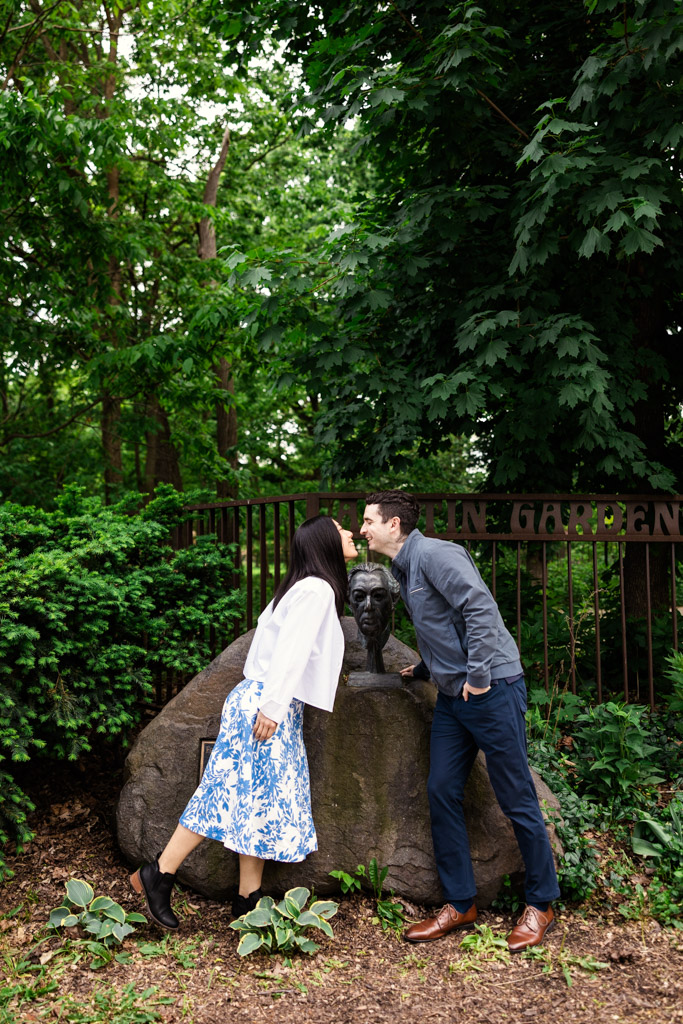 Engaged couple leans in to kiss in front of a bronze bust in Austin Gardens in Oak Park, Illinois