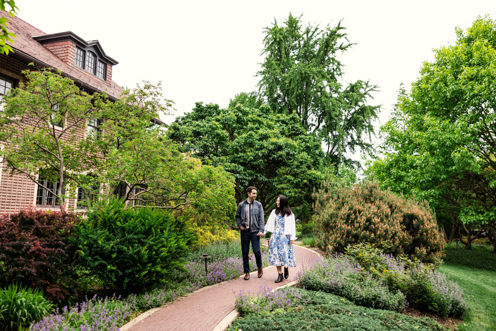 A couple walks hand in hand on a garden path surrounded by lush greenery and flowering plants outside Cheney Mansion during their Oak Park engagement session