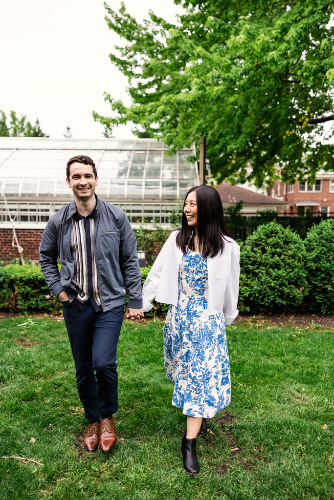 A man and woman walk together near Cheney Mansion on grass, smiling, with lush greenery and a greenhouse in the background during their Oak Park engagement session