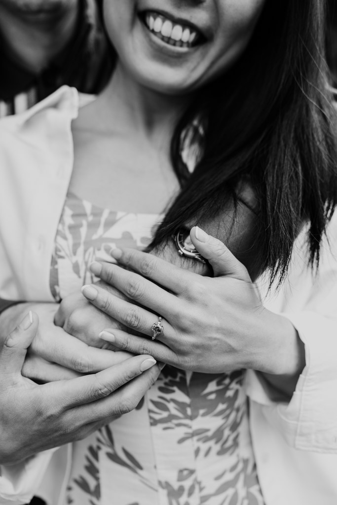Black and white photo of woman smiles as her partner embraces her , showing an engagement ring on her finger during Oak Park engagement session