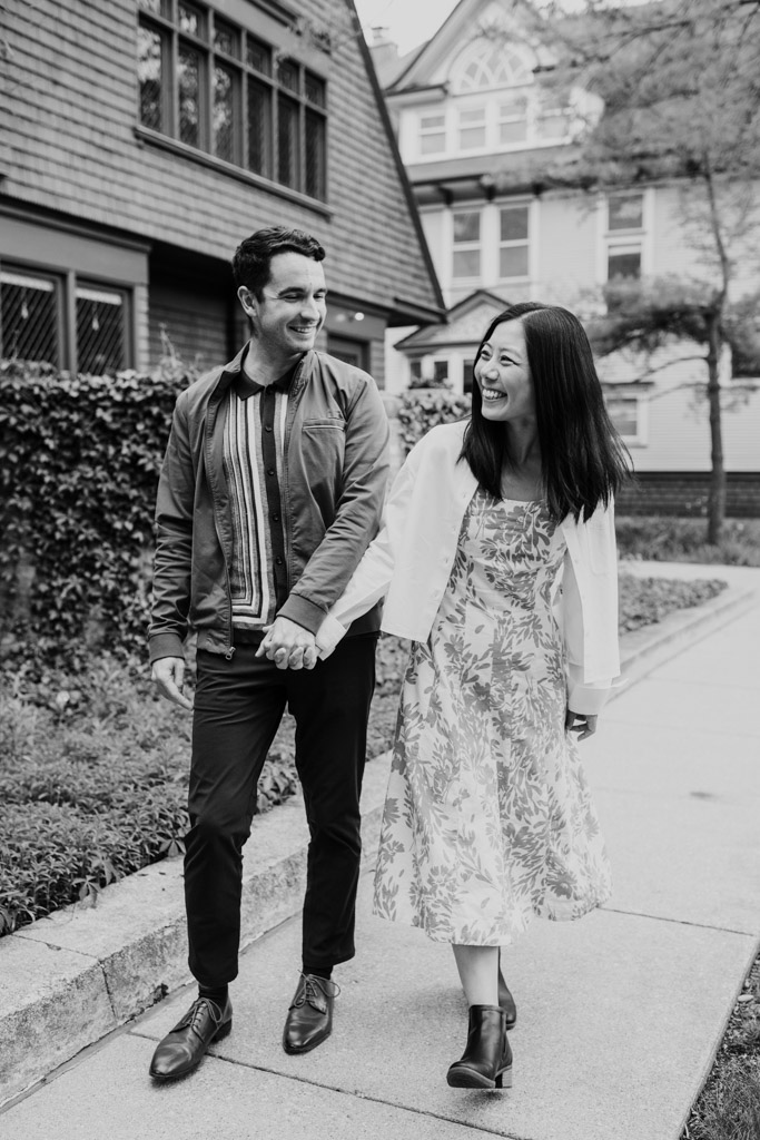 Black and white photo of smiling couple walking hand in hand on a sidewalk in front of houses, looking at each other during their Oak Park engagement session