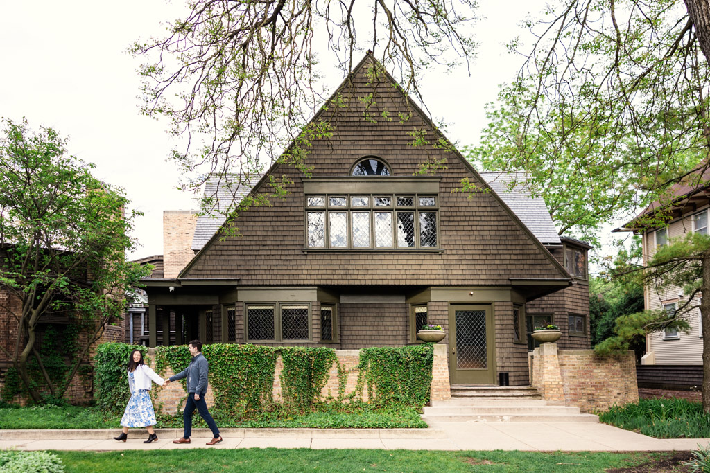 Engaged couple holding hands walks past the Frank Lloyd Wright Home and Studio in Oak Park, Illinois, a brown, ivy-covered house with large windows and a steep roof