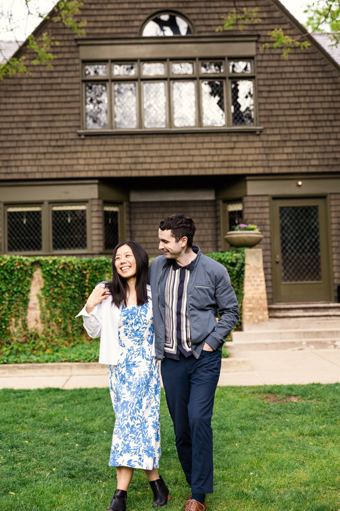 A smiling couple walks arm-in-arm on grass in front of the Frank Lloyd Wright Home and Studio during their Oak Park engagement session