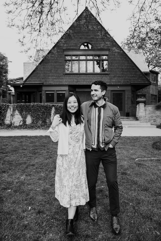 Black and white portrait of smiling couple stands on a lawn in front of the Frank Lloyd Wright Home and Studio during their Oak Park engagement session
