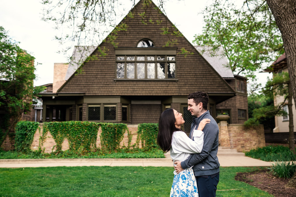 Couple embraces and smiles at each other outside the Frank Lloyd Wright Home and Studio with greenery and large windows during their Oak Park engagement session