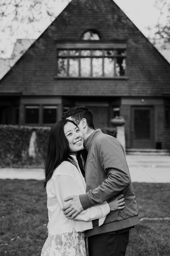 Black and white photo of man whispering in his partner's ear as they stand in front of the Frank Lloyd Wright Home and Studio during their Oak Park engagement session