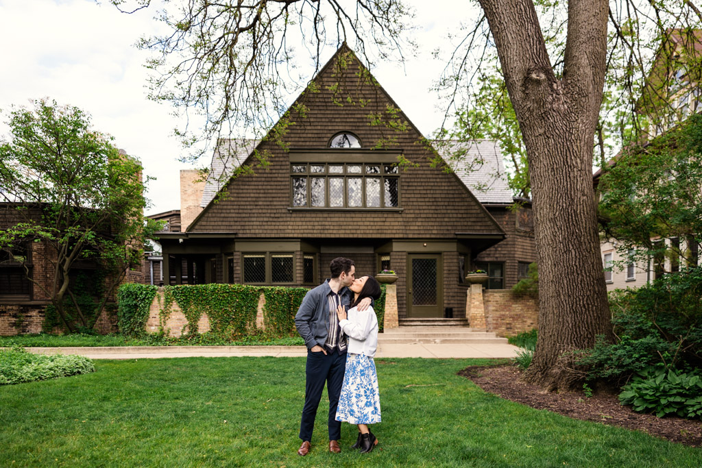 A couple stands close together, kissing in front of a large, brown house with green lawn and trees—capturing their Oak Park engagement.