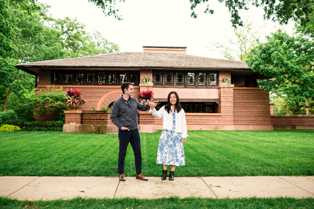 Engaged man and woman stand smiling on a lawn in front of the Frank Lloyd Wright Home and Studio in Oak Park