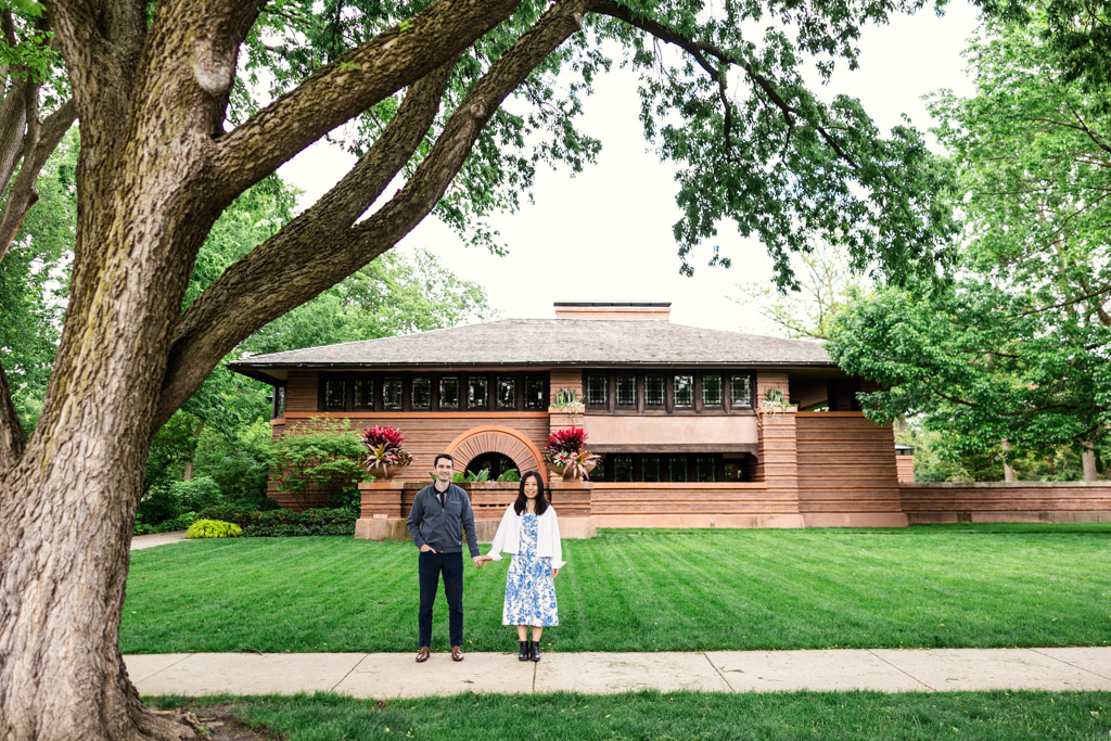 Engaged couple stands on a sidewalk in front of the Frank Lloyd Wright Home and Studio in Oak Park with a lush green lawn and trees