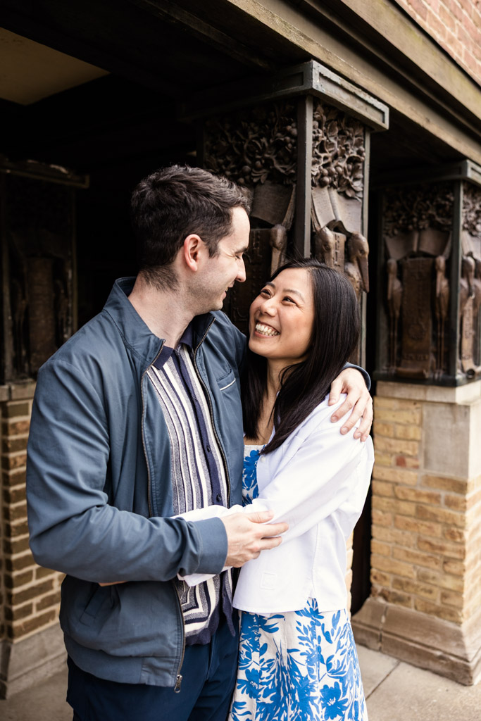 A couple smiles and embraces during their Oak Park engagement session outside the Frank Lloyd Wright Home and Studio with detailed carvings and brick walls