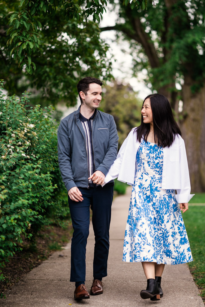 Couple walks hand in hand on a path in Oak Park, smiling at each other and surrounded by greenery during their spring engagement session