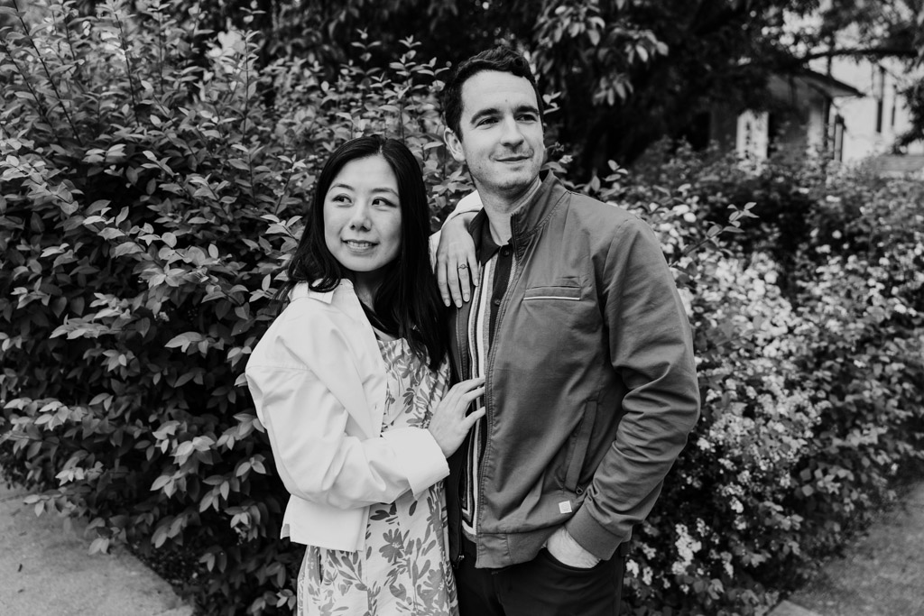 Black and white portrait of woman and man standing close together near a row of bushes and trees in a residential Oak Park neighborhood
