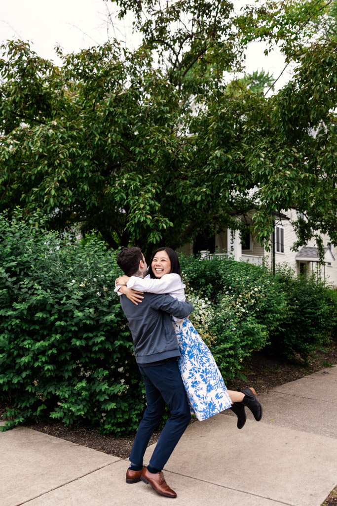 Man lifts his smiling partner in a white and blue dress on a sidewalk by green bushes during their Oak Park engagement session