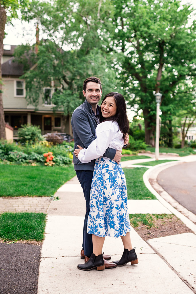 A smiling couple hugs on a sidewalk in a leafy suburban neighborhood during their Oak Park engagement session
