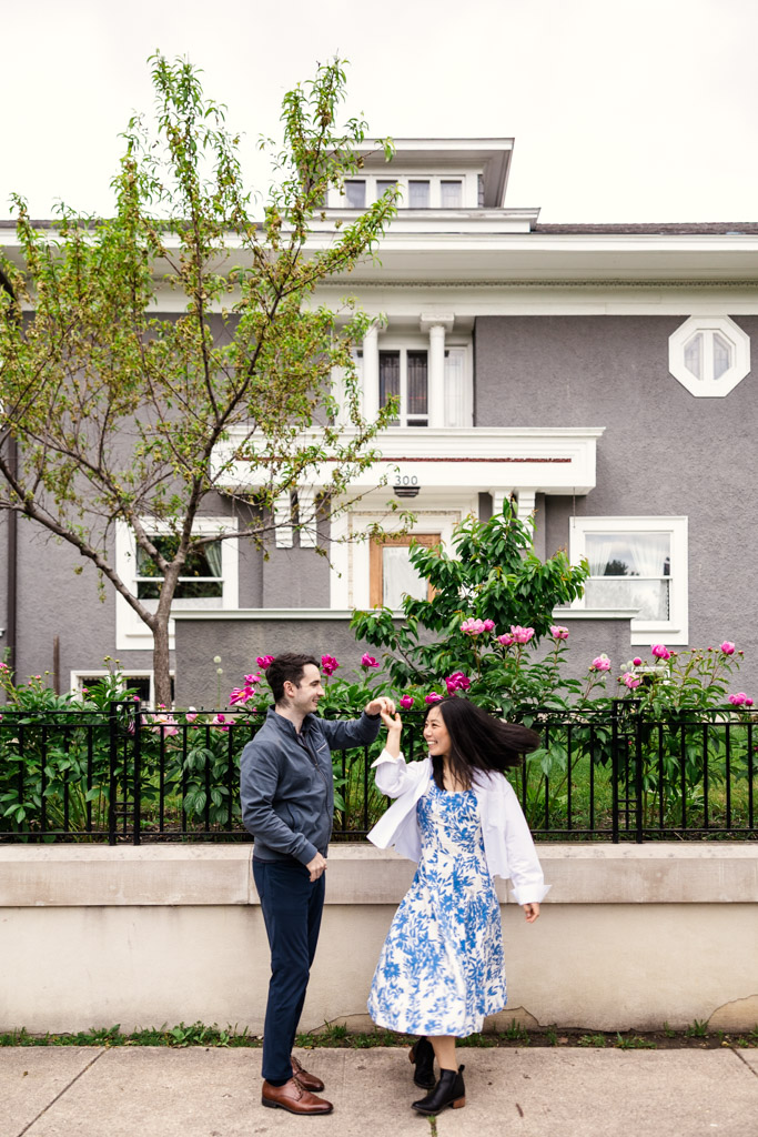 A couple dances on the sidewalk in front of a gray house with blooming flowers and a small tree during their Oak Park engagement session