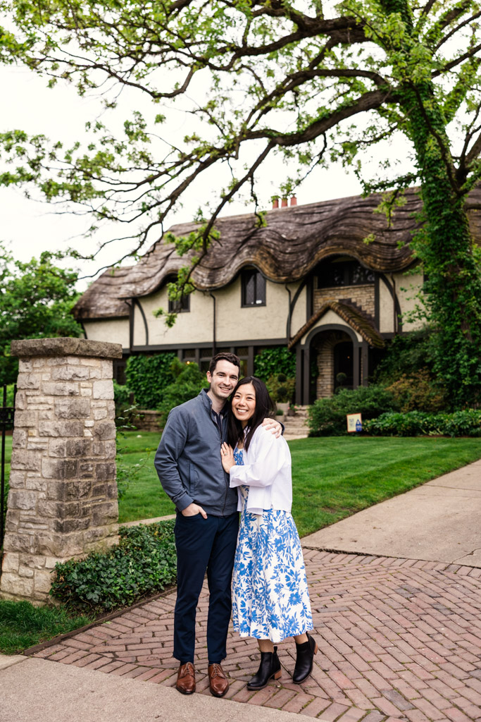 Portrait of smiling couple stands together in front of a charming, thatched-roof house with lush greenery during their Oak Park engagement session