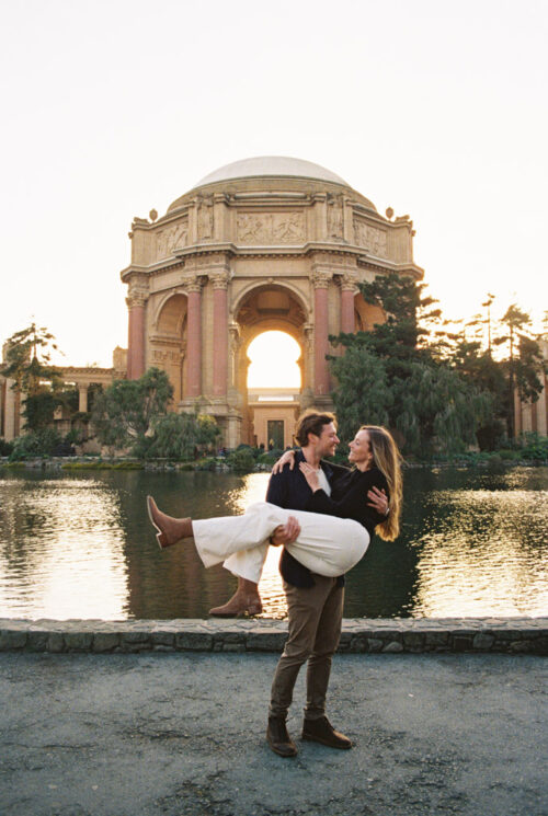 Film photo of man holding his fiancée by a pond, with the Palace of Fine Arts in the background during San Francisco engagement session