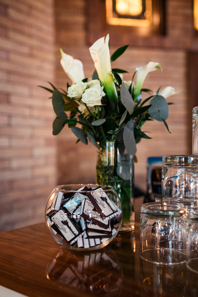 A vase of white flowers and a glass bowl of custom matchboxes sit on a wooden counter with stacked glasses for Cafe Brauer wedding reception