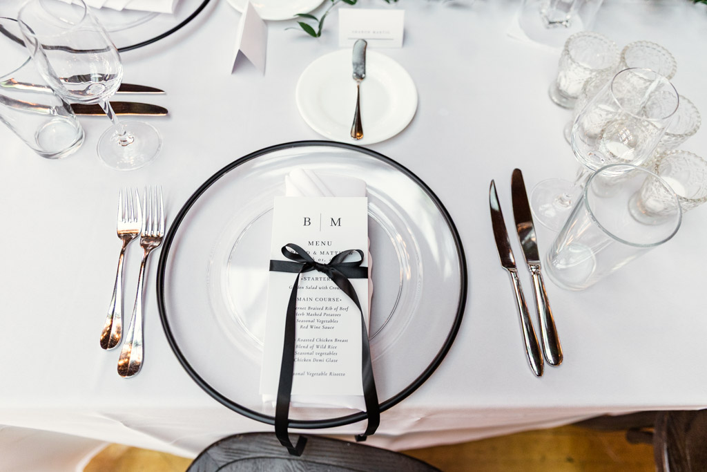 Guest place setting for Cafe Brauer wedding reception with a clear plate, black-ribboned menu, silver cutlery, and glassware on a white tablecloth