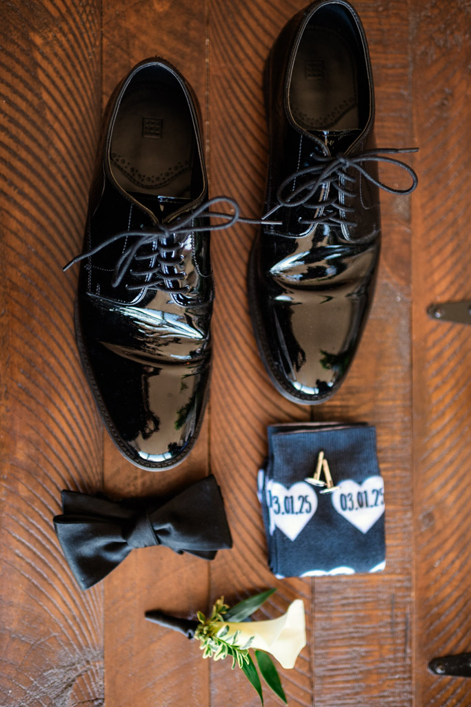 Groom's black dress shoes, black bow tie, boutonniere, and custom patterned socks arranged on a wooden surface