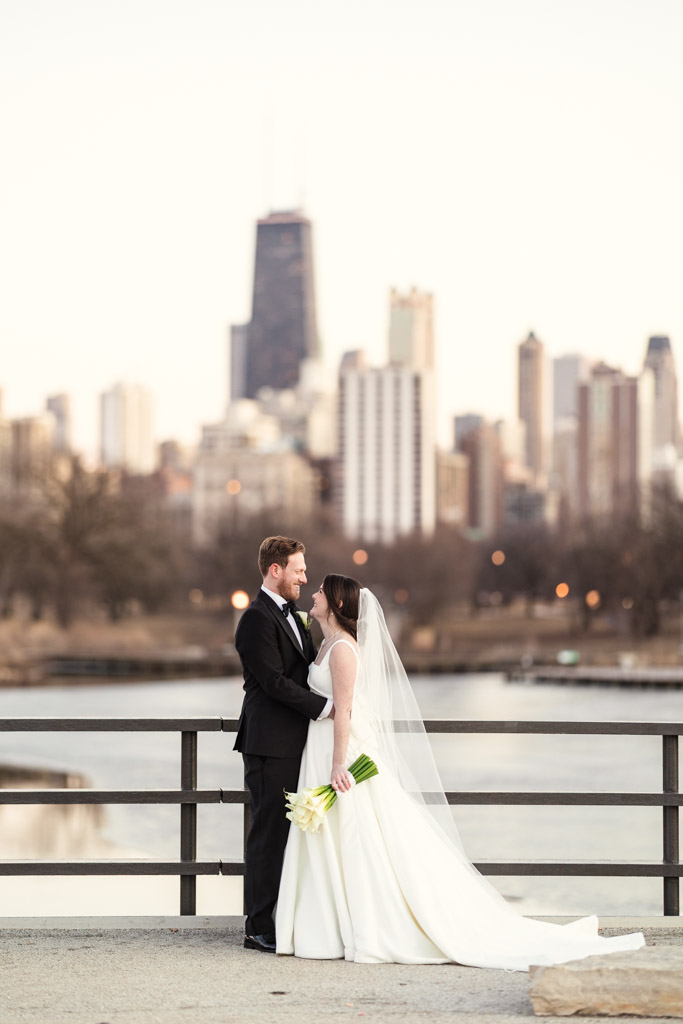 Bride and groom in wedding attire stand together on a Lincoln Park bridge near Cafe Brauer, with the Chicago skyline in the background