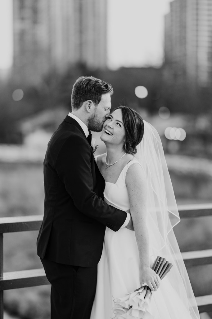 Black and white photo of groom kissing his smiling bride on the cheek as they pose together on a Lincoln Park bridge near Cafe Brauer