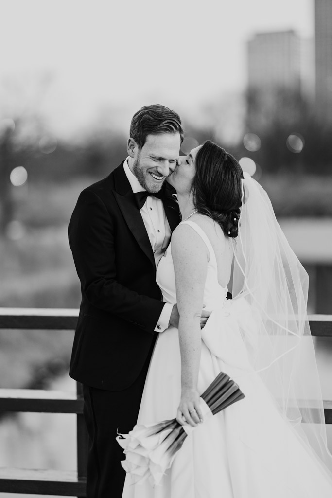 Black and white photo of bride kissing groom on the cheek while holding her bouquet on a Lincoln Park bridge near their Cafe Brauer wedding reception