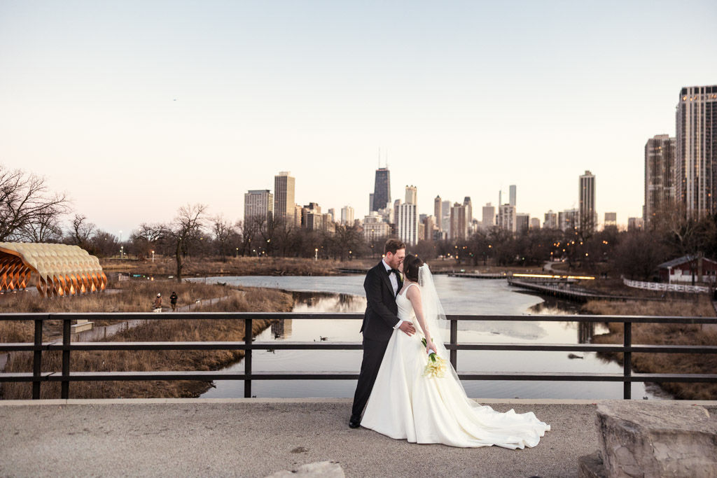 Bride and groom embrace on a Lincoln Park bridge near Cafe Brauer, with the Chicago skyline and the Honeycomb background at sunset