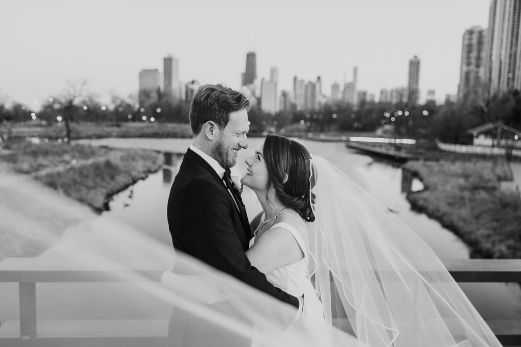 Creative black and white portrait of bride and groom holding each other and smiling on a bridge in Lincoln Park with the bride's flowing veil and the Chicago skyline in the background